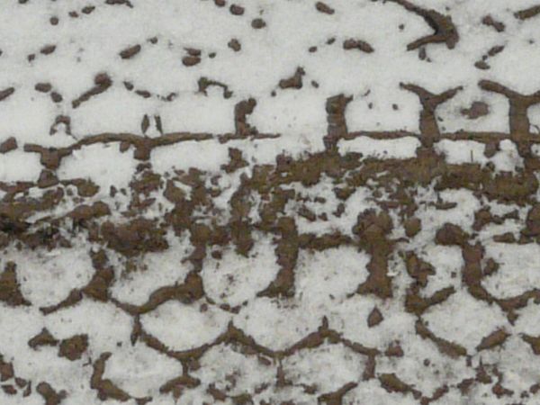 Dirt road texture in dark brown tone with tire marks and thin layer of snow across surface.